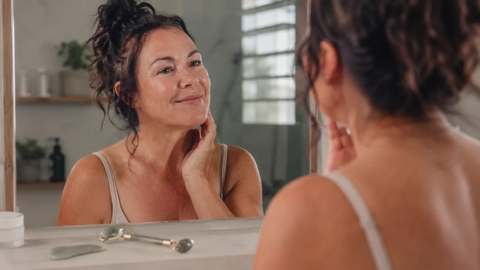 Woman smiling at her reflection after her morning gua sha routine, tools resting on the counter beside her