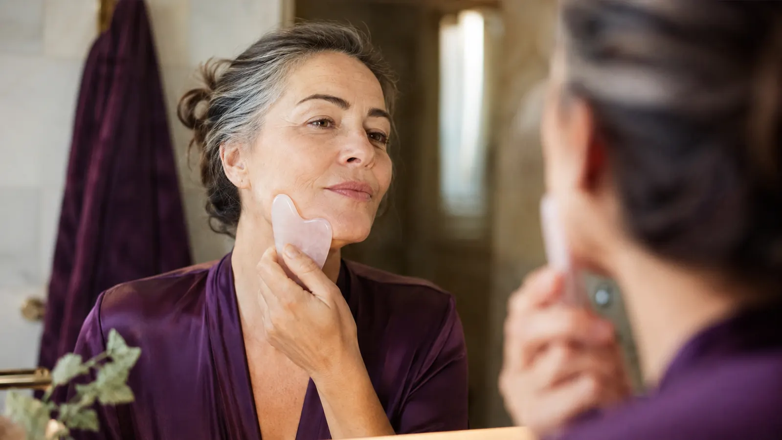 Woman in her fifties holding a rose quartz gua sha stone to her jawline in warm morning bathroom light