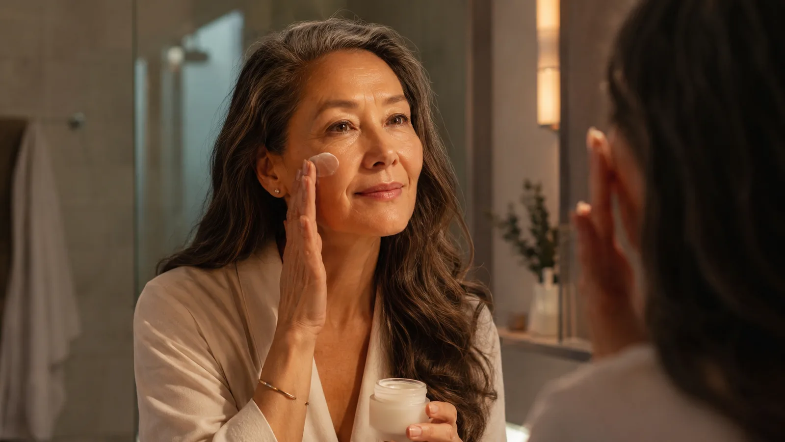 Woman in her 50s smiling at her reflection in a softly lit bathroom mirror after applying retinol cream