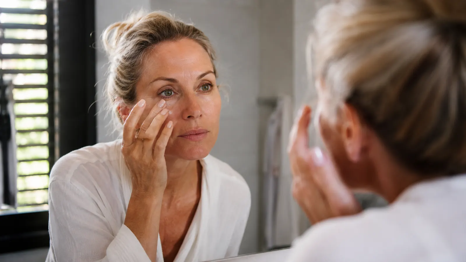Woman in her 50s applying eye cream with her ring finger in front of a bathroom mirror in soft morning light