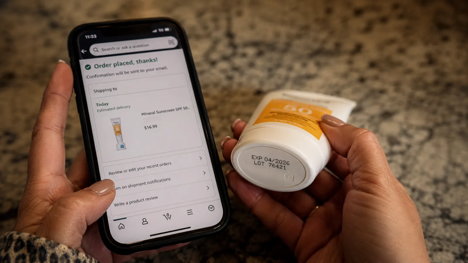 Close up of hands checking a sunscreen tube's expiration date next to a phone showing an Amazon order screen