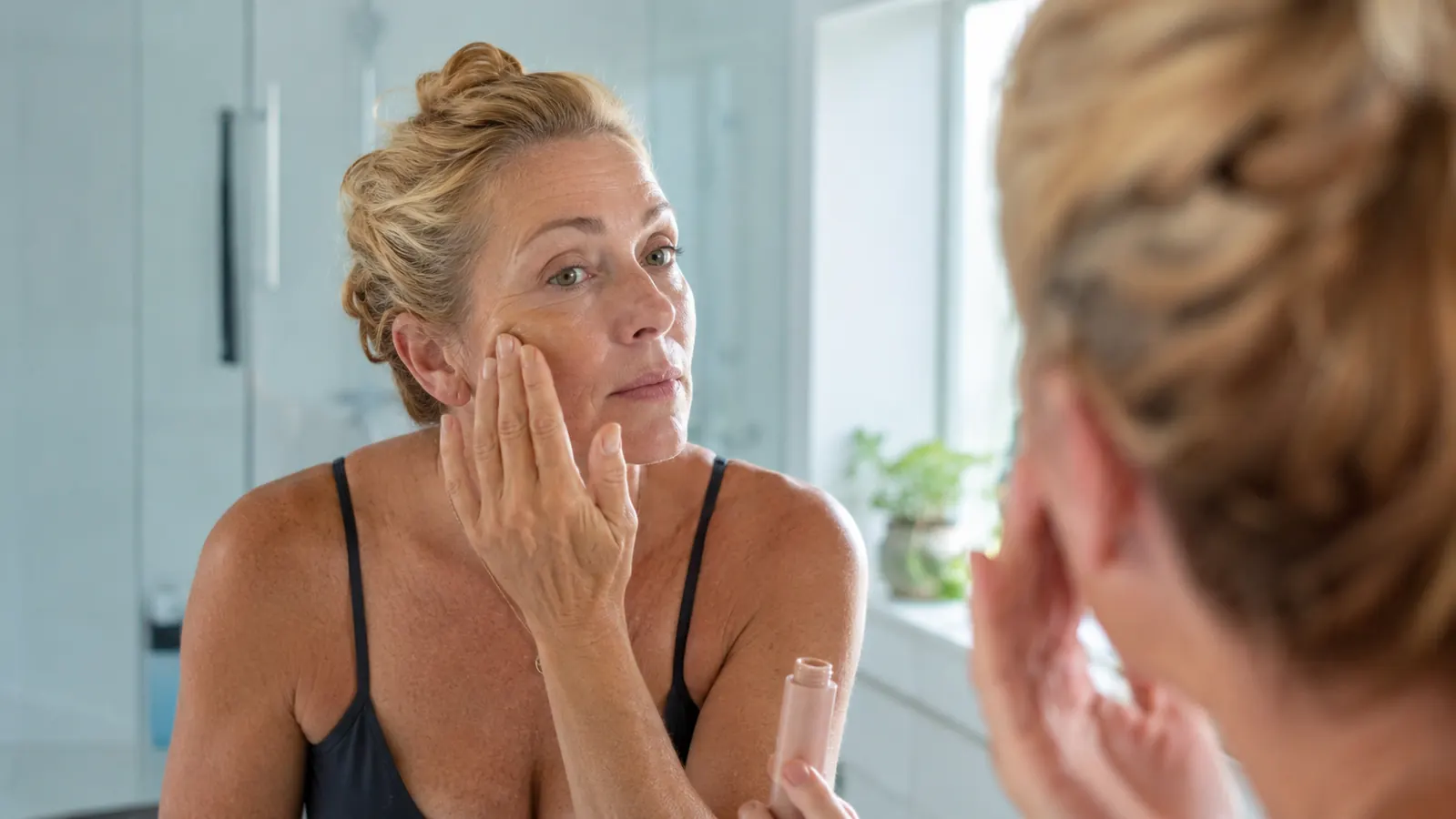 Woman applying tinted mineral sunscreen at a bathroom mirror in honest, unfiltered morning light