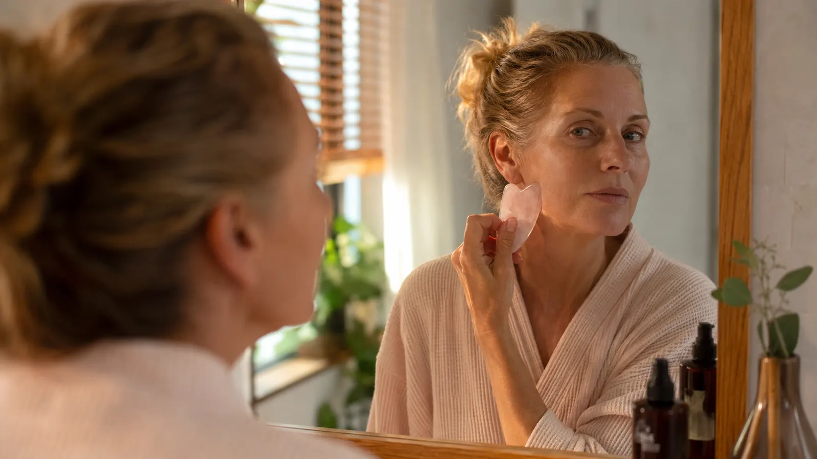 Woman in her fifties standing at a sunlit bathroom mirror holding a rose quartz gua sha stone to her jawline
