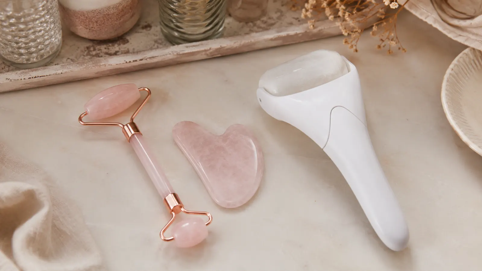 Rose quartz roller and gua sha stone laid side by side on a vanity next to a plain plastic ice roller