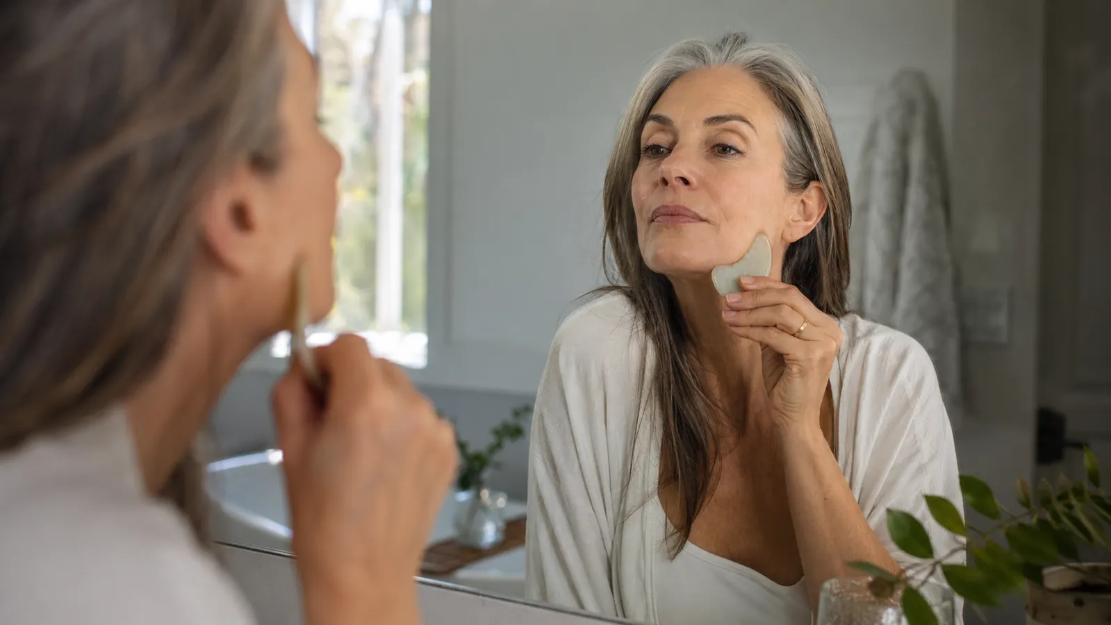 Woman in her 50s using a gua sha stone along her jawline in a bright bathroom mirror in the morning