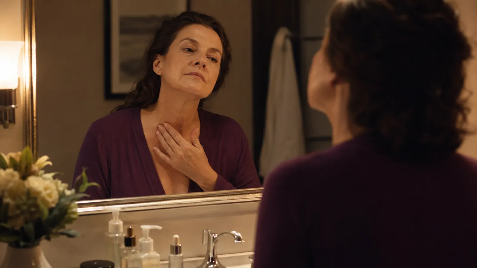 Woman in her fifties at a bathroom mirror in soft evening light, one hand resting at the base of her neck, checking her sagging skin before her nightly routine
