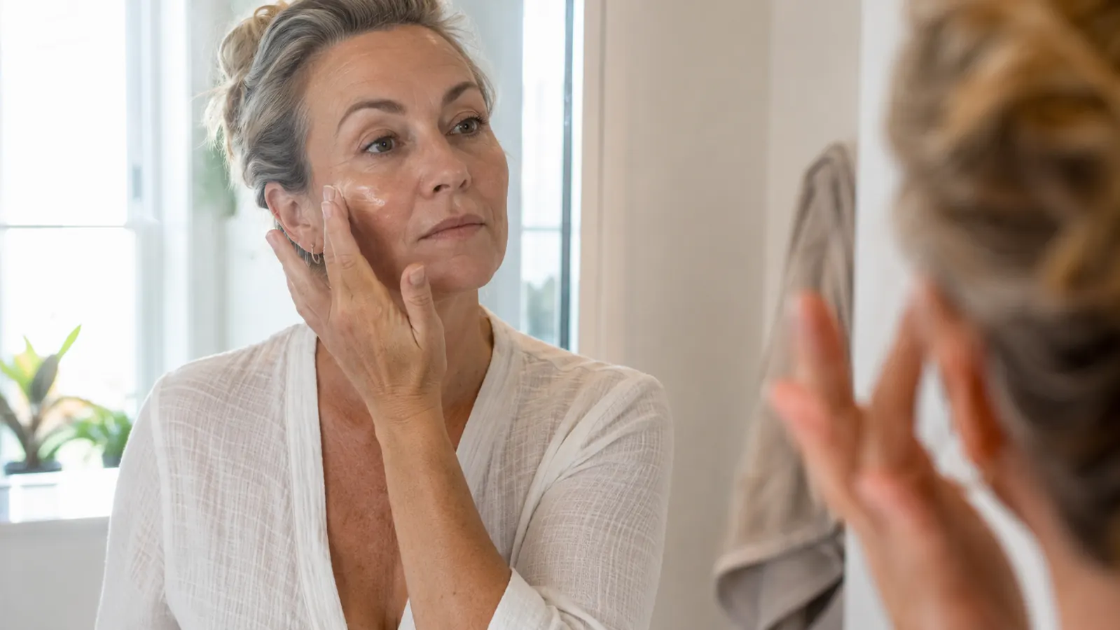 Woman in her 50s applying sunscreen to her face in natural morning light near a sunny window
