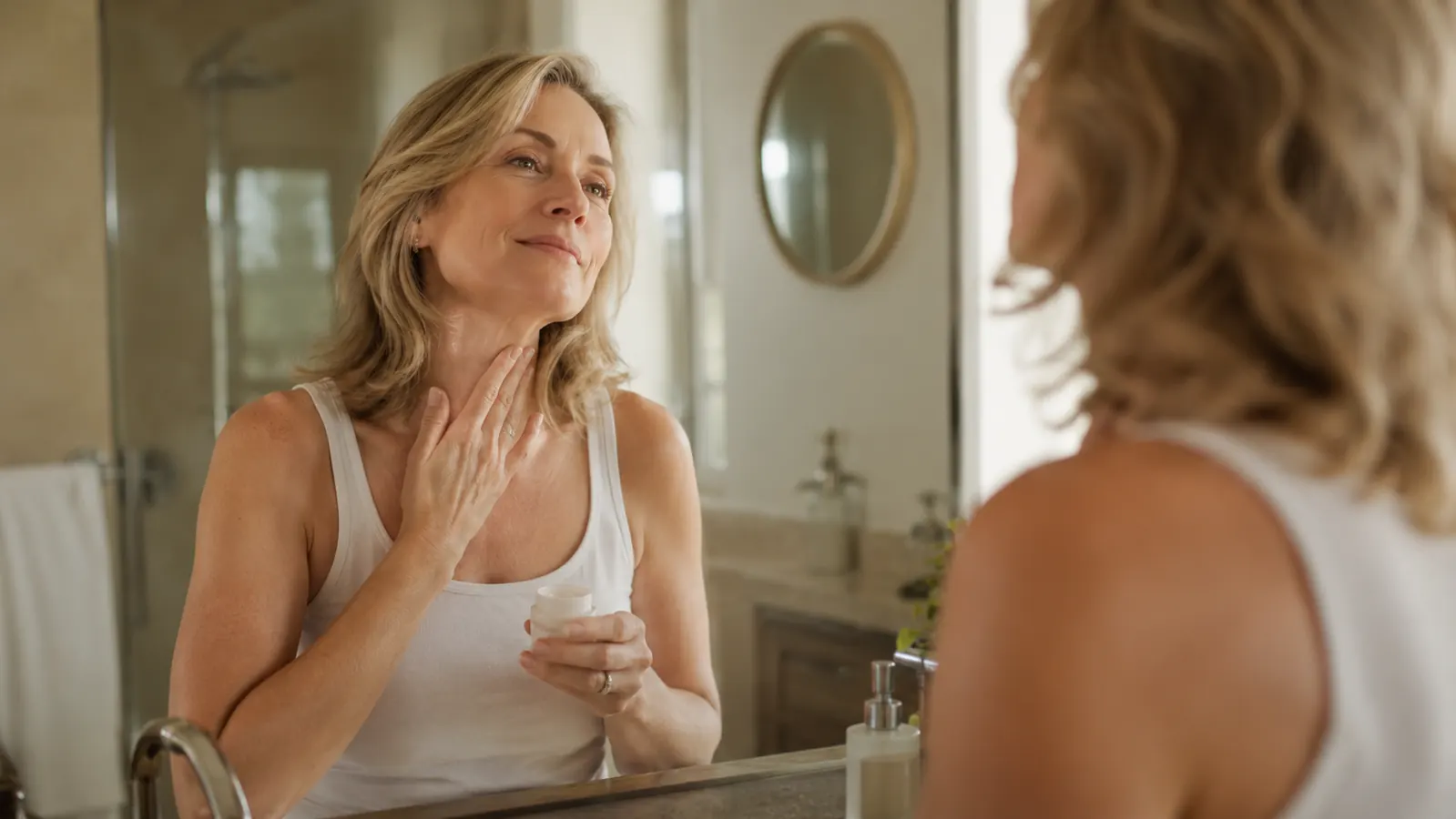 Woman in her mid fifties applying cream to her neck in a warm bathroom mirror, soft morning light