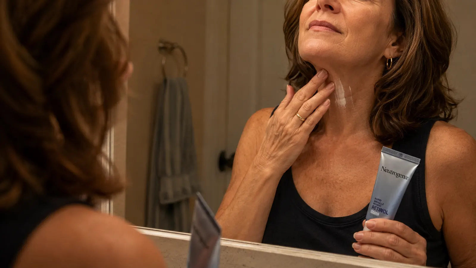 Hand smoothing a pea sized dot of retinol cream from the jawline down onto the neck in front of a bathroom mirror