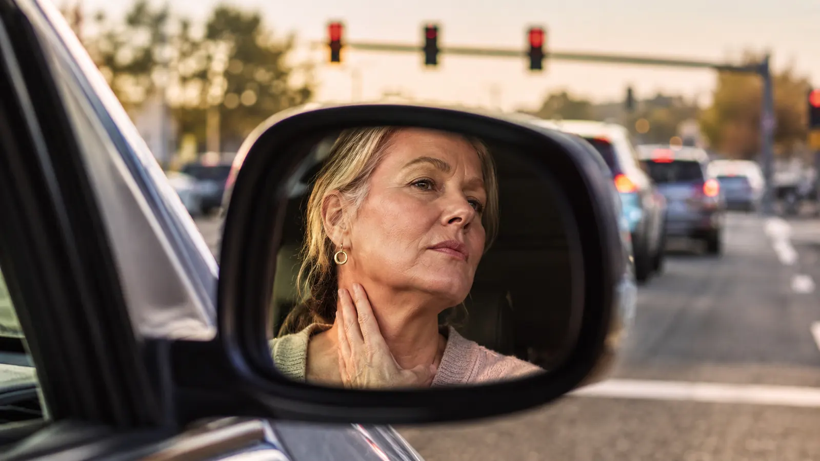 Woman in her early 50s glancing at her neck in a car side mirror at a red light, soft afternoon light